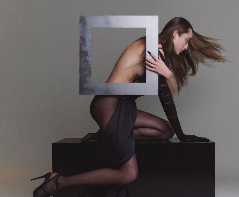 Adult female model in sheer tights, gloves, and high heels kneeling on black cube, framed by geometric metallic art piece in photo studio.