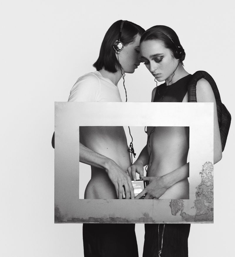 Two women wearing headphones stand in front of a framed black and white photo of torsos holding a music player in a minimalist style.