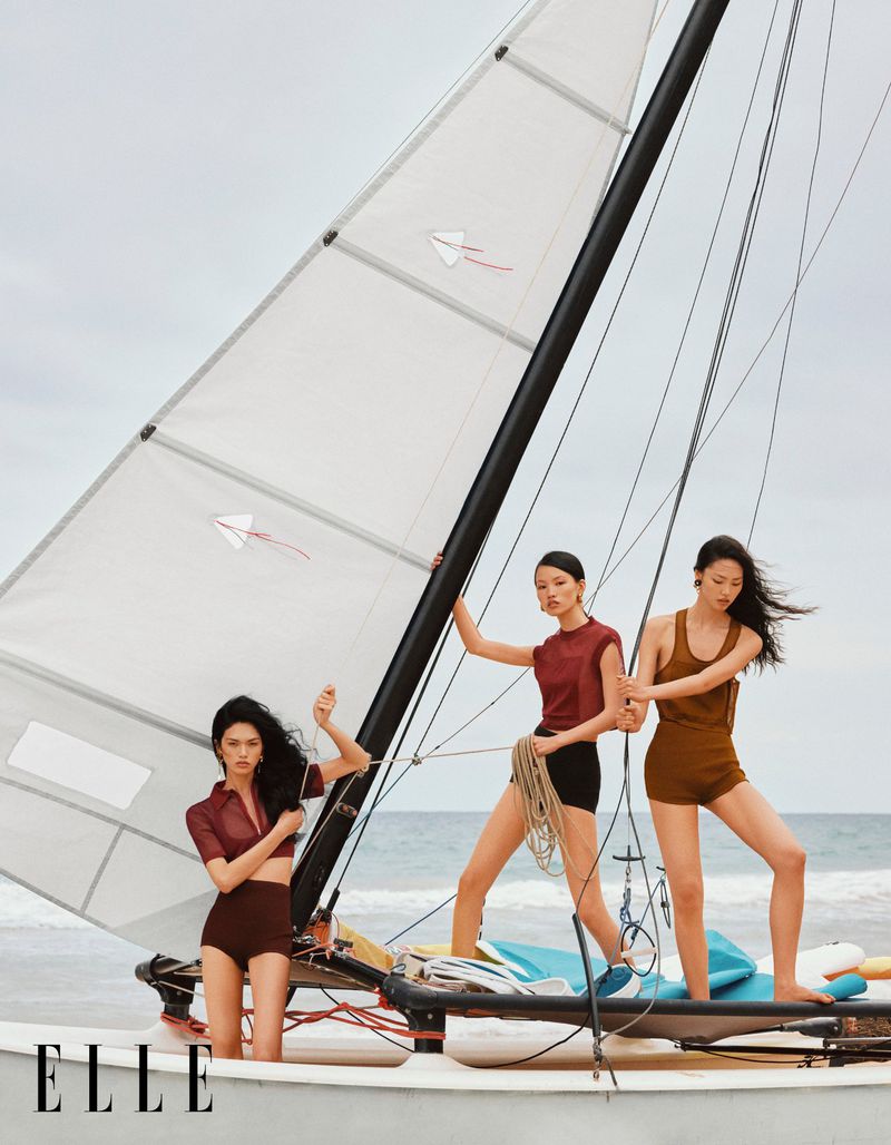 Three Asian female fashion models pose on a sailboat on a beach in an editorial fashion shot for ELLE magazine in the daytime with a cloudy sky.