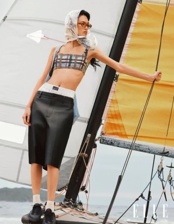 An adult female fashion model is posing on a sailboat, wearing a scarf, a plaid bra top and black shorts near the ocean.