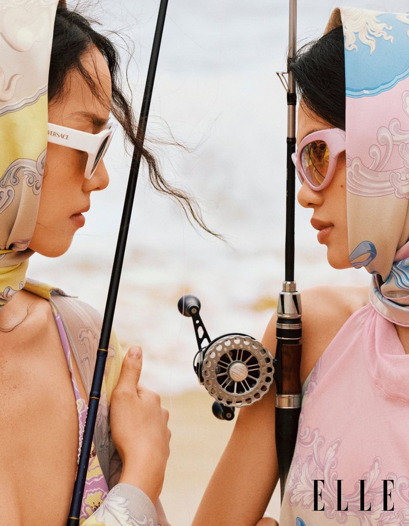 Two women in headscarves and sunglasses pose with fishing poles on a beach in a fashion editorial, with ocean and sky background.