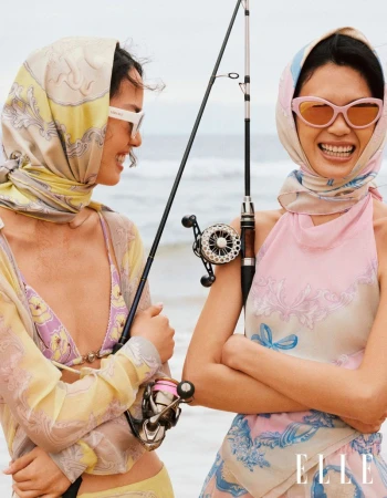 Two women in headscarves and sunglasses pose with fishing rods on a beach under overcast skies; fashion editorial with pastel colors.