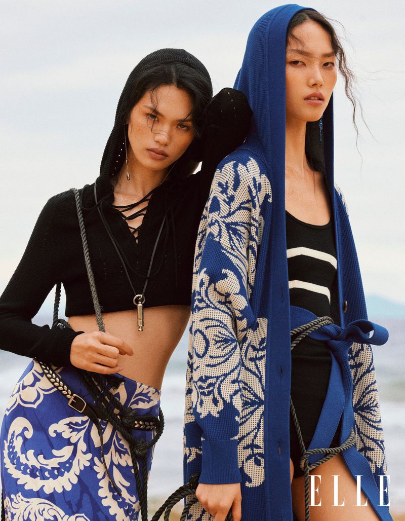 Two Asian women pose outdoors wearing hooded cardigans and patterned skirts in a fashion editorial, showcasing knitwear designs against a muted backdrop.