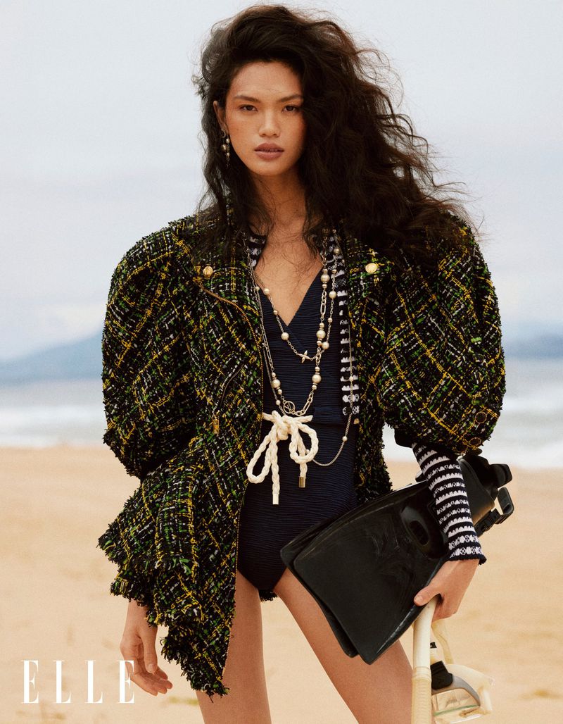 Editorial fashion shot of an Asian female model on a sandy beach wearing a black swimsuit, green jacket, and holding snorkeling gear.