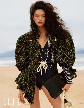 Editorial fashion shot of an Asian female model on a sandy beach wearing a black swimsuit, green jacket, and holding snorkeling gear.