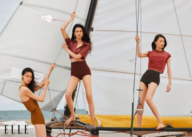 Three Asian female models on a sailboat, wearing high-waisted shorts and matching tops, posing in a fashion editorial on the water.