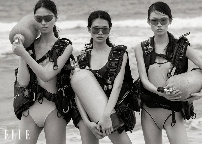 Three Asian women in swimwear and scuba gear pose on a beach with dive tanks in a black and white fashion shot.