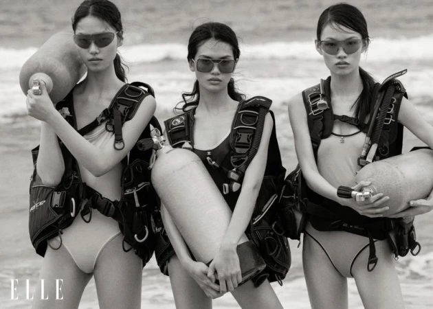 Three Asian women in swimwear and scuba gear pose on a beach with dive tanks in a black and white fashion shot.