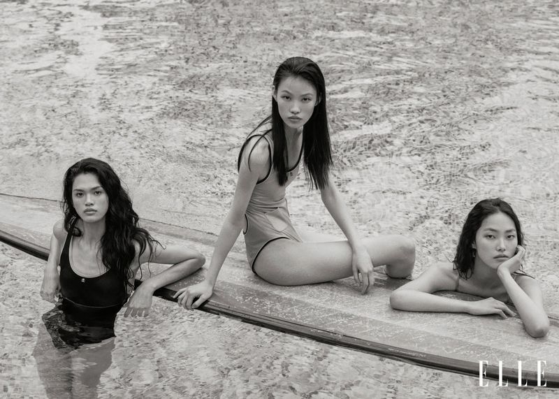 Monochrome shot of three Asian adult females in swimsuits, posing in water by a pool edge, Elle logo visible, soft natural lighting.