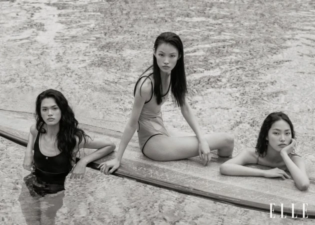 Monochrome shot of three Asian adult females in swimsuits, posing in water by a pool edge, Elle logo visible, soft natural lighting.