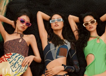 Three female models wearing colourful swimwear and sunglasses recline together on a dark textured net surface.