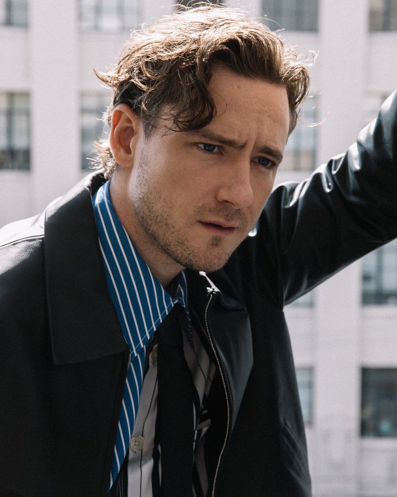Portrait of a man with dark blonde hair wearing a striped blue and white shirt and a black jacket in an urban setting.
