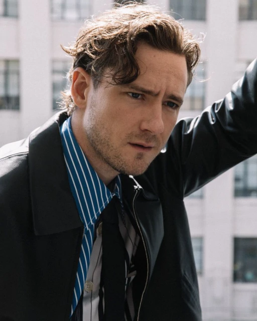 Portrait of a man with dark blonde hair wearing a striped blue and white shirt and a black jacket in an urban setting.