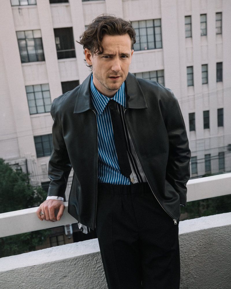 Adult male posing on a rooftop with a city building in the background, wearing a black leather jacket, and a vertically striped blue shirt.
