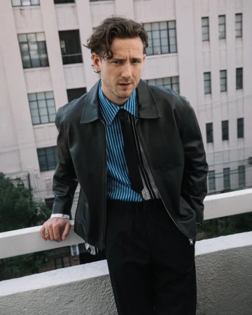 Lewis Pullman wearing a dark leather jacket and striped shirt standing outdoors against a building exterior backdrop