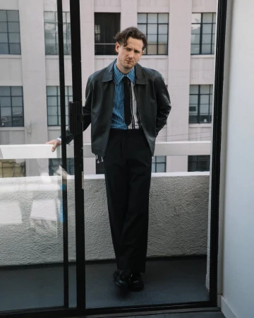 Actor Lewis Pullman leaning on a balcony railing wearing a black leather jacket and striped shirt for Numero Netherlands.