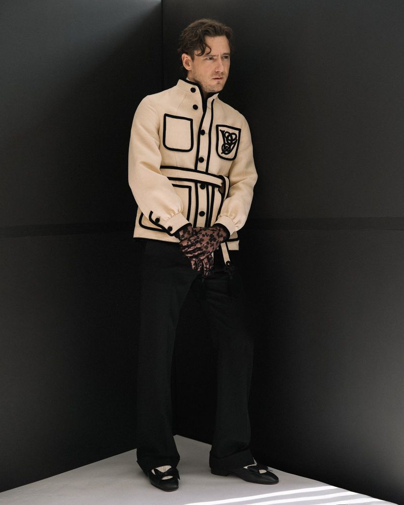 Actor Lewis Pullman wearing a cream jacket with black piping and dark trousers standing in a dark studio corner.