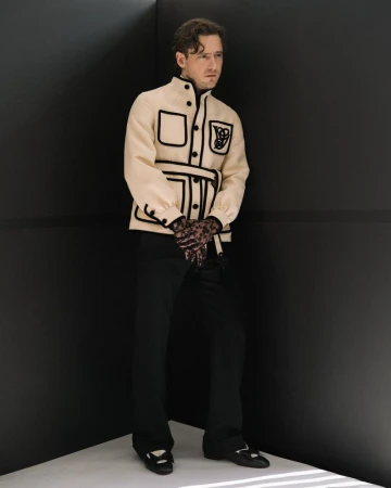 Actor Lewis Pullman wearing a cream jacket with black piping and dark trousers standing in a dark studio corner.