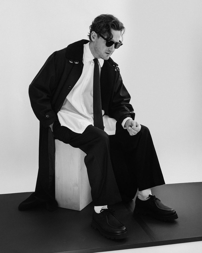 Stylish adult male in a coat and tie sitting on a wooden box in a studio, creating a sophisticated and modern fashion portrait.