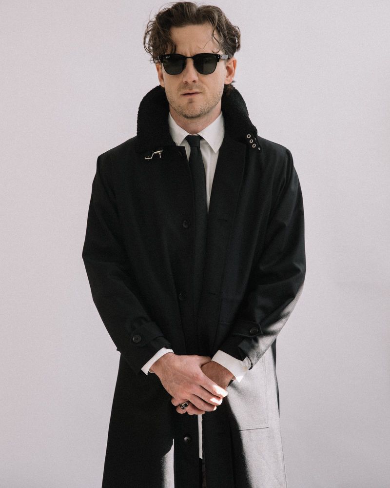 Actor Lewis Pullman wearing dark sunglasses and a black coat over a white shirt with a tie against a white background