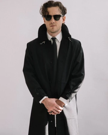 Actor Lewis Pullman wearing dark sunglasses and a black coat over a white shirt with a tie against a white background