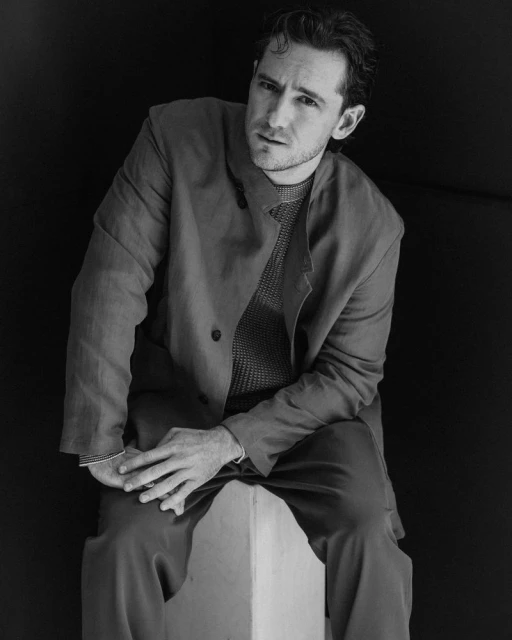 Monochrome portrait of a man in an open blazer sitting on a white cube against a black background, looking at the camera.