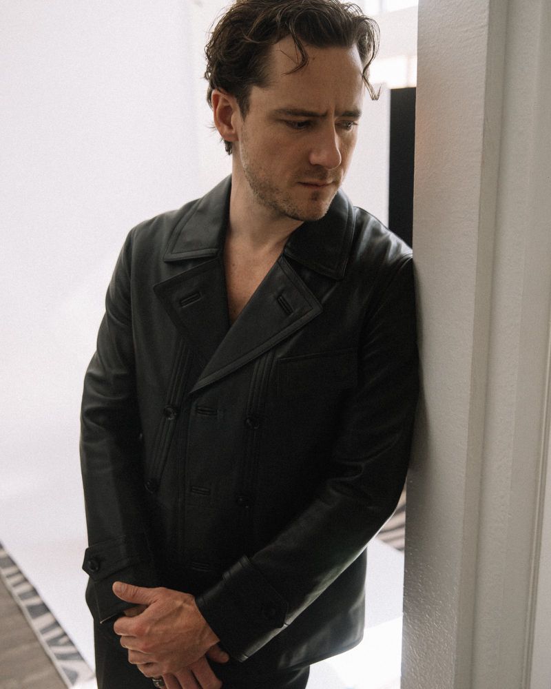 Portrait of a serious man with dark hair wearing a black leather jacket in a studio setting against a white backdrop with bright lighting.