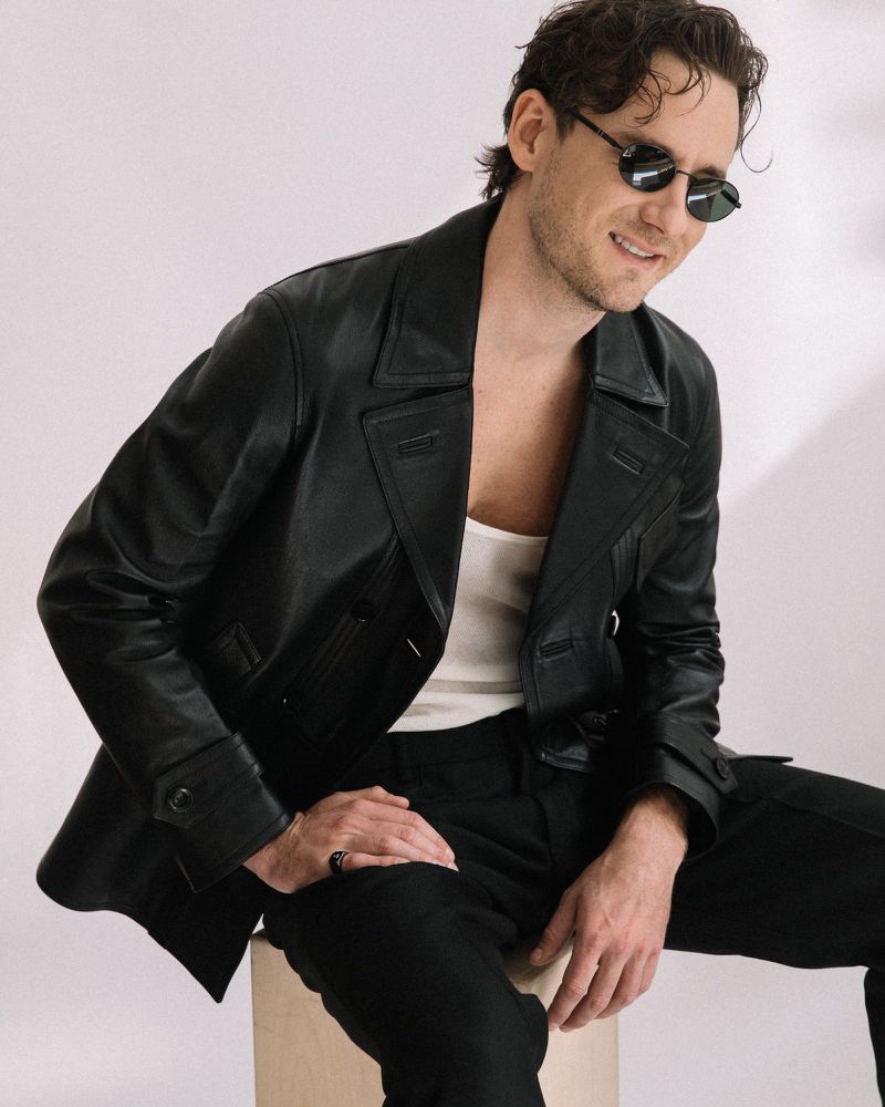 Lewis Pullman wearing a black leather jacket and sunglasses seated against a plain white background for Numero Netherlands.