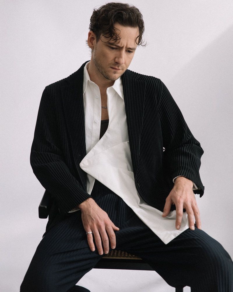 A man sits on a chair, wearing a black corduroy suit over a white shirt, looking down against a plain white background.