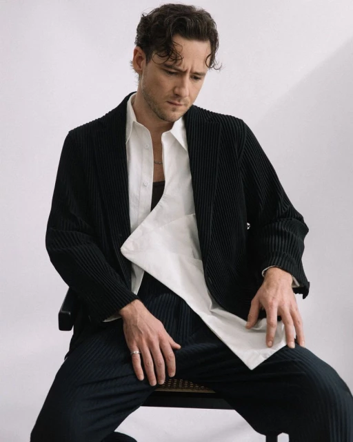 A man sits on a chair, wearing a black corduroy suit over a white shirt, looking down against a plain white background.