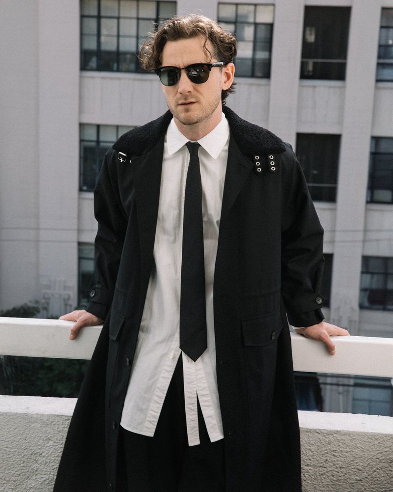 Man in a black trench coat, white shirt, and black tie stands on a balcony with a cityscape background, showcasing fashion and style.