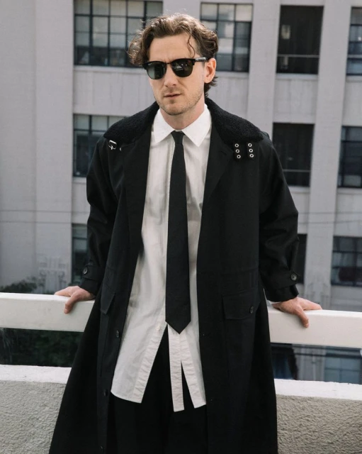 Man in a black trench coat, white shirt, and black tie stands on a balcony with a cityscape background, showcasing fashion and style.