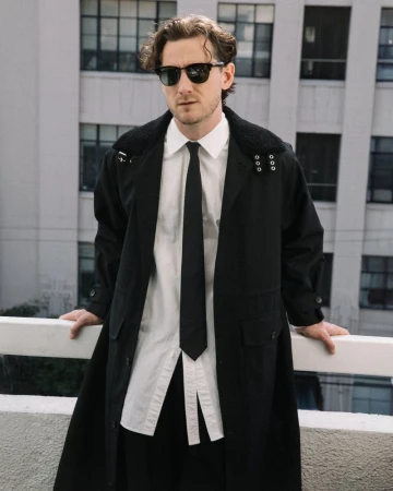 Man in a black trench coat, white shirt, and black tie stands on a balcony with a cityscape background, showcasing fashion and style.