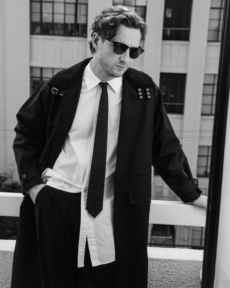 Portrait of Lewis Pullman wearing sunglasses, a dark coat, and a tie, standing against an urban building background.