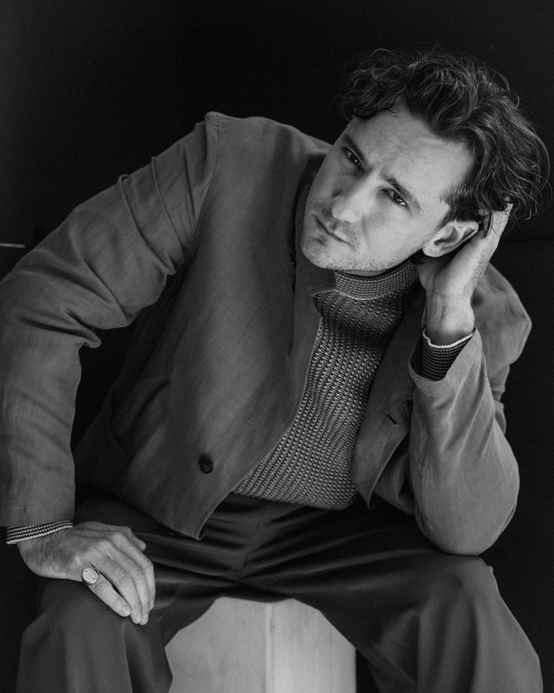 Monochrome portrait of a man in a suit and turtleneck sweater sitting indoors with hand behind head, looking off-camera with neutral expression.