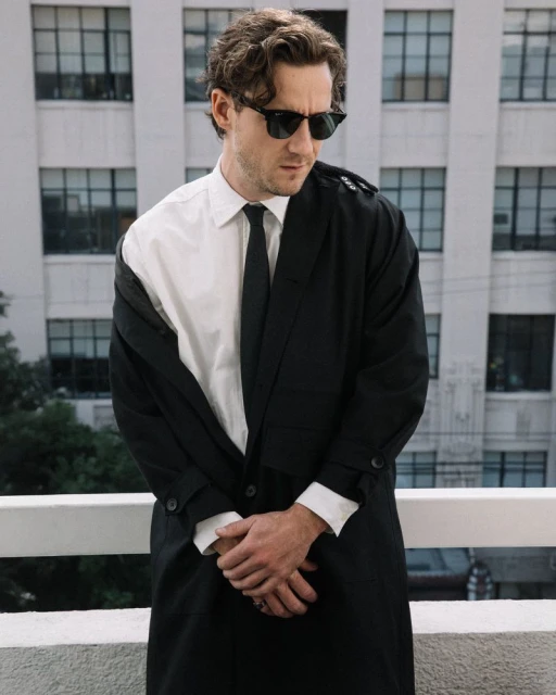 Man in black and white coat and sunglasses stands on balcony with city building backdrop. Fashion editorial style, medium shot.
