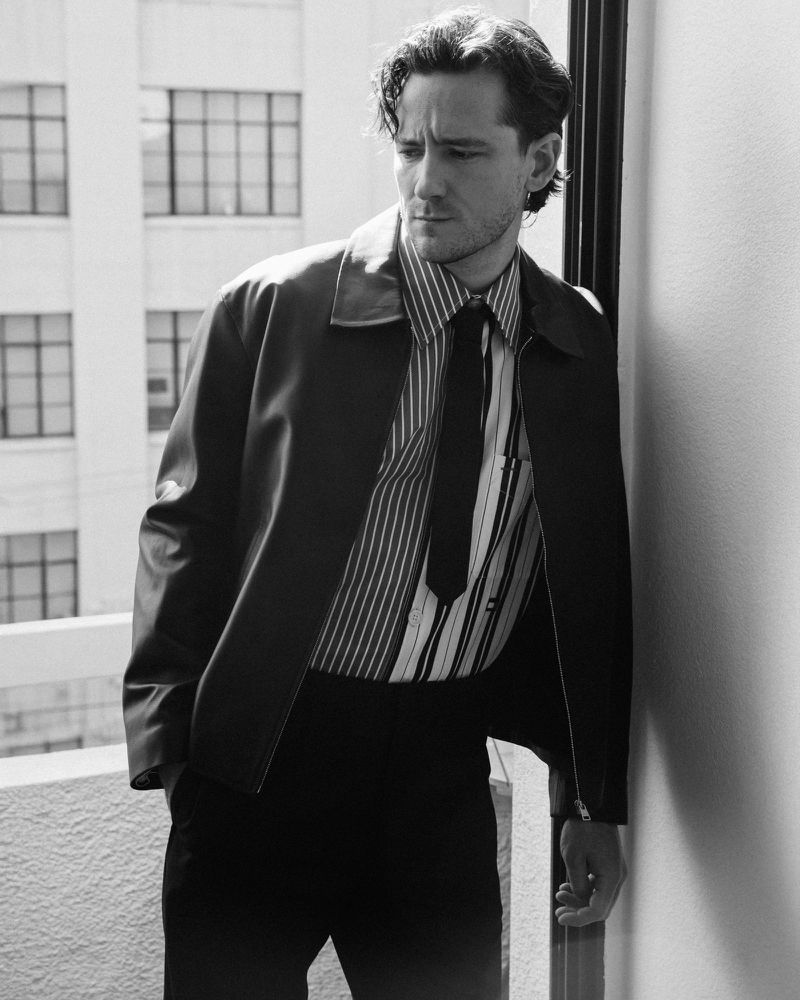 Portrait of Lewis Pullman wearing a striped shirt and dark jacket standing on a balcony for Numero Netherlands