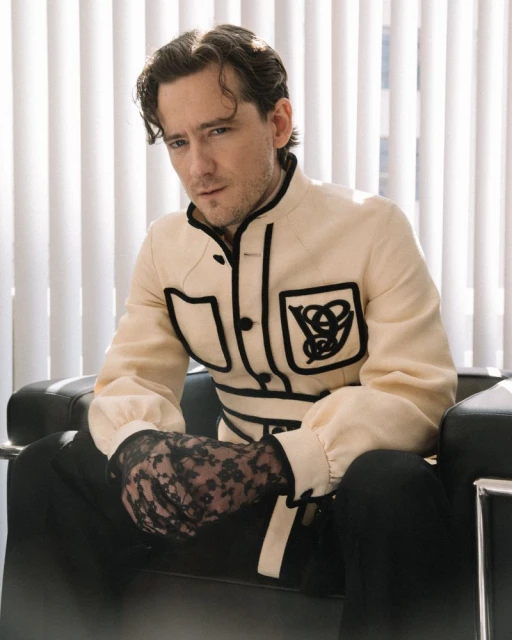 Portrait of an adult man in a cream-colored jacket with black trim and black lace gloves, seated against a background of white blinds.