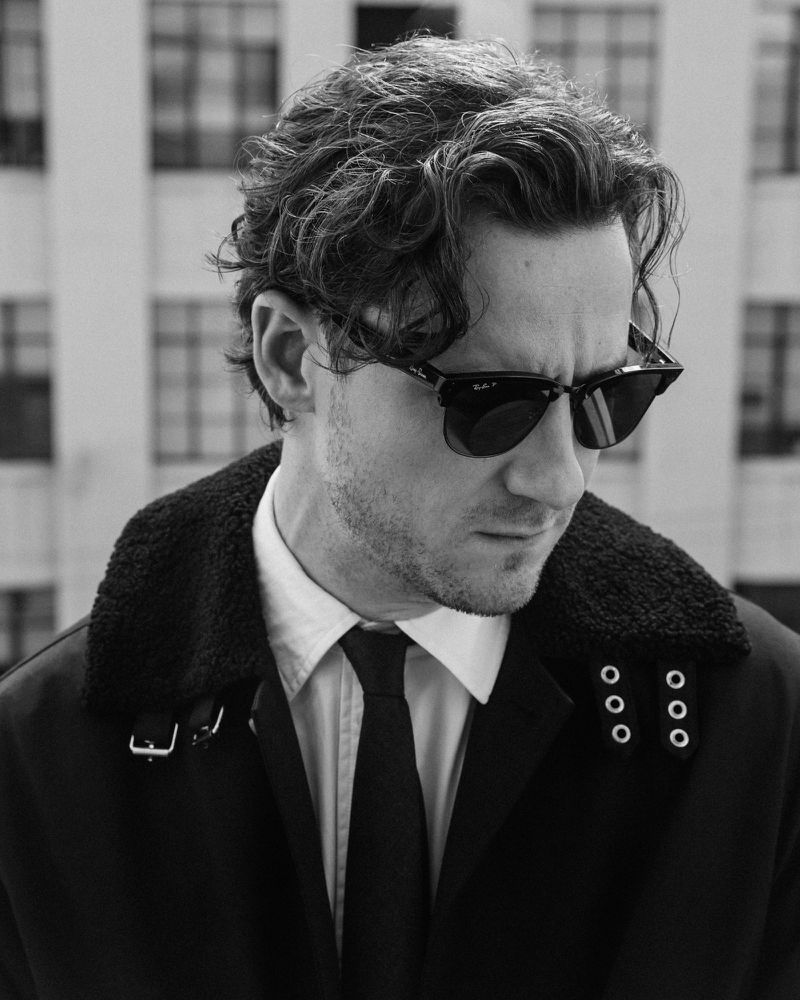 Black and white portrait of a man wearing sunglasses, a white collared shirt, and a dark jacket against a light-colored building.