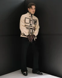 Actor Lewis Pullman wearing a cream jacket with black piping and dark trousers standing in a dark studio corner.