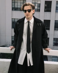 Actor Lewis Pullman wearing sunglasses, white shirt, black tie, and long black coat leaning on a white railing outdoors