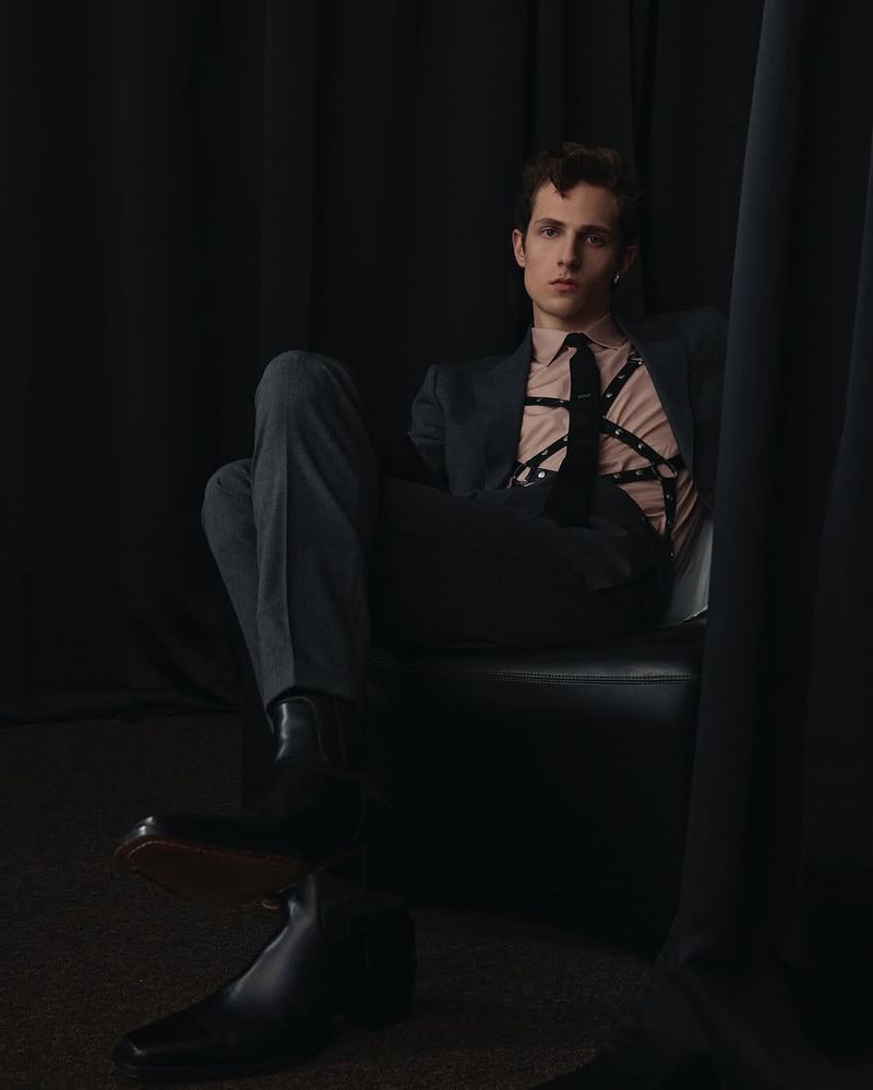Portrait of a slender adult male in a suit, leather harness, and boots, sitting indoors against a dark curtain backdrop in a studio setting.