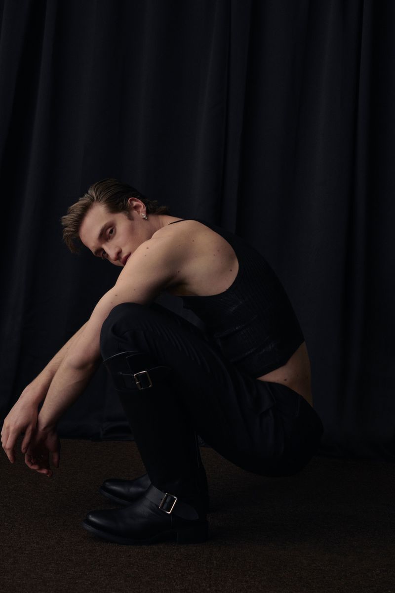 Young male fashion model in black tank top, dark pants, and boots posing in a photo studio against a black curtain backdrop.