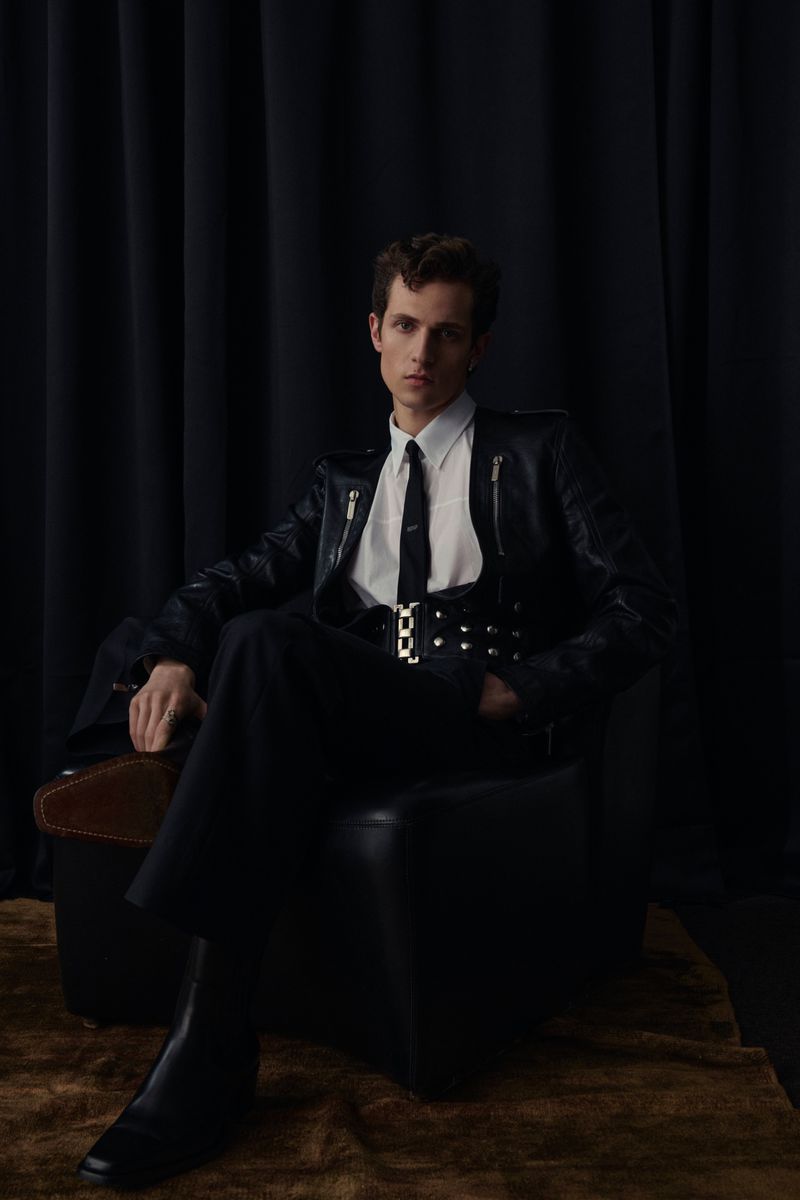 A seated male model wearing a black leather jacket, white shirt, and tie against dark curtains for Man About Town.