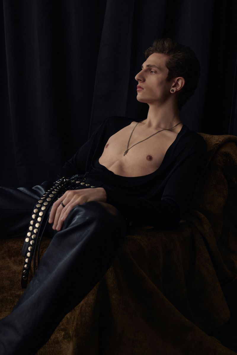 Portrait of a slender man in dark clothing reclining on a couch, looking off to the right side with a moody and dramatic vibe.
