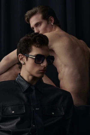 Two adult men in a dark studio setting. One man is wearing sunglasses and a black leather jacket with a tie, the other is shirtless.