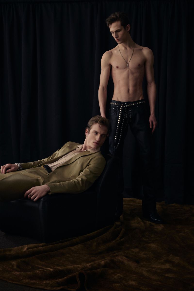 Two male fashion models pose in a dimly lit studio with a black backdrop and brown shag carpet, one seated in a black chair.
