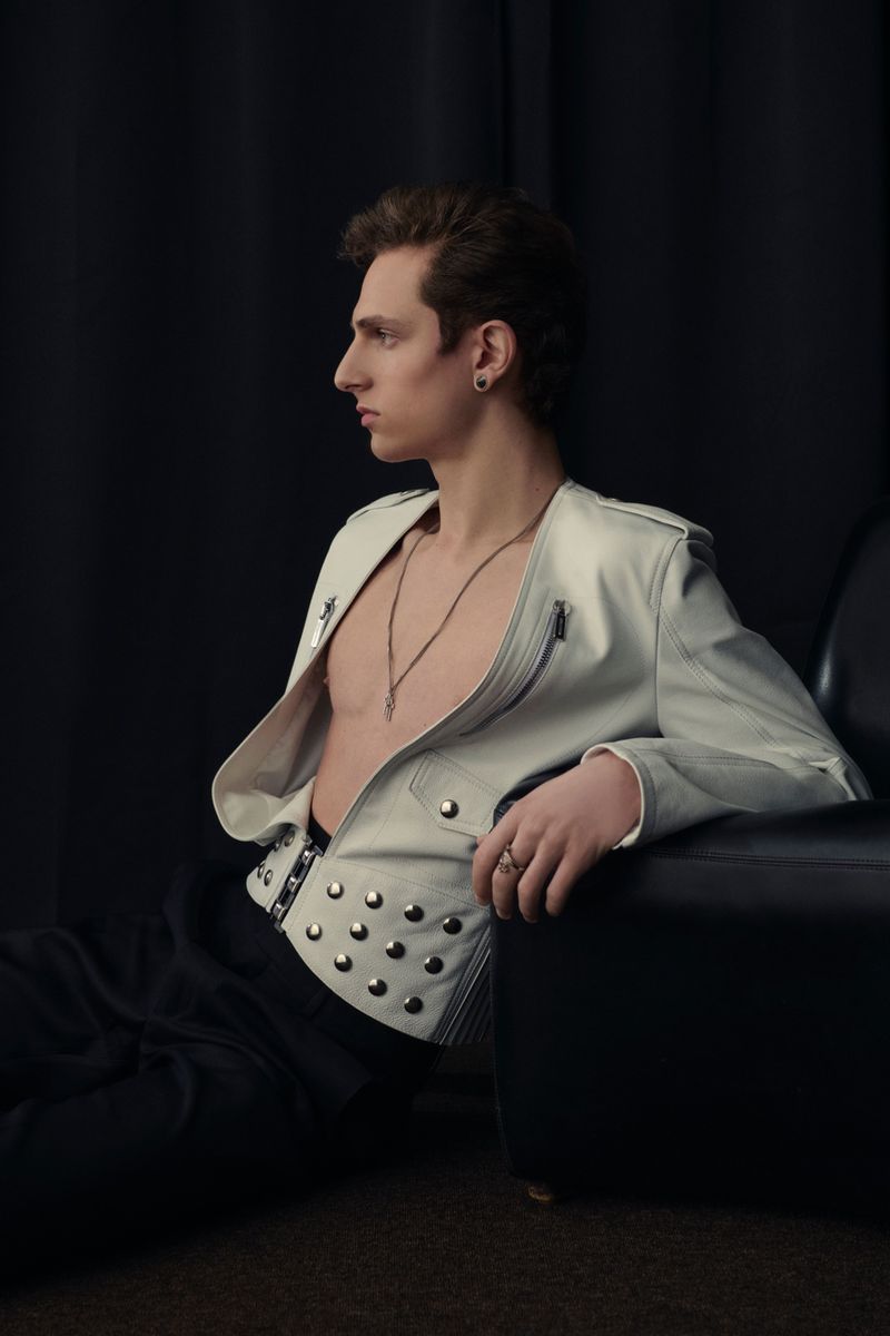 Profile shot of a young man wearing a white jacket with studs, seated in front of a black backdrop and next to a black chair.