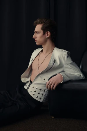 Profile shot of a young man wearing a white jacket with studs, seated in front of a black backdrop and next to a black chair.