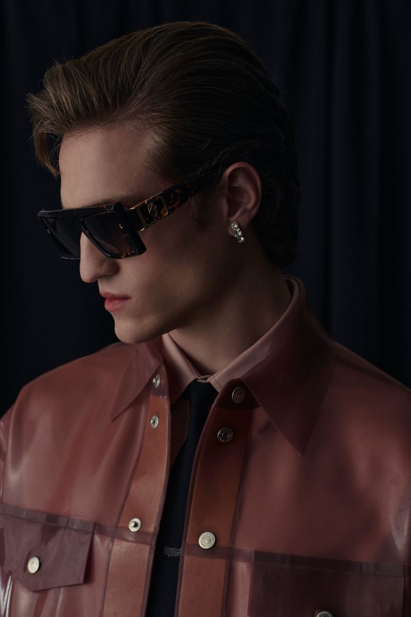 Close-up studio shot of a young adult male wearing sunglasses and a pink translucent jacket, against a dark background.